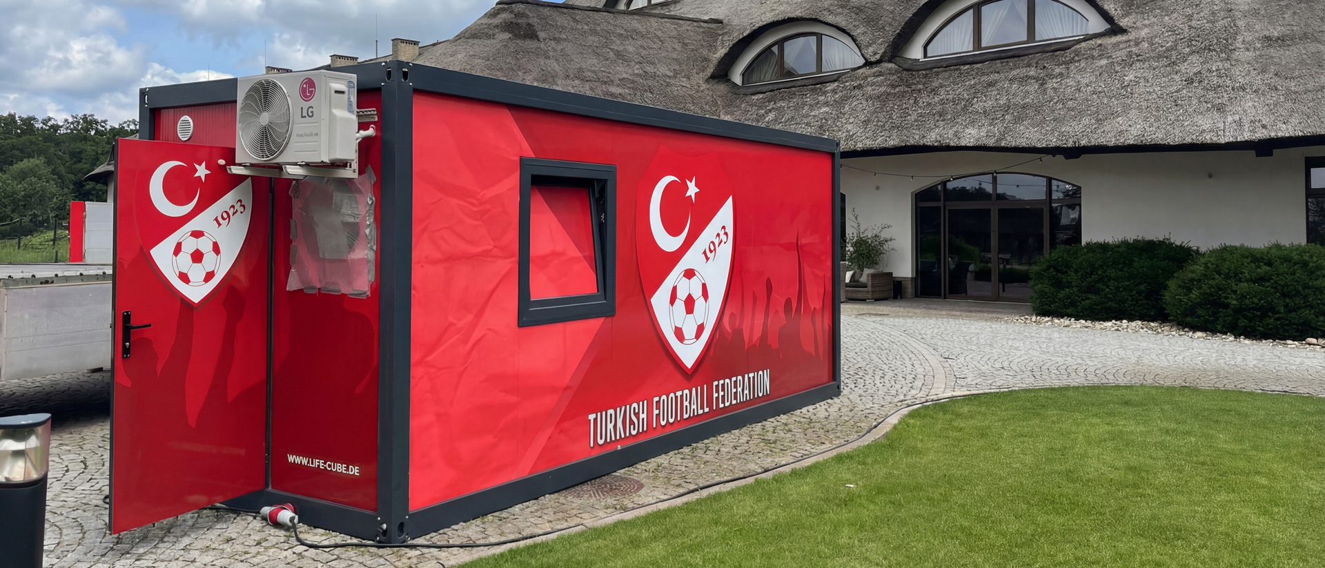 Ein roter LifeCube Container mit Logos des türkischen Fußballverbands steht auf einem gepflasterten Vorplatz.